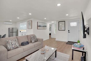 A modern living room with a beige sofa, a marble coffee table, and a small side table with books and a plant. - Boardwalk Bungalow 1  For 6 (Kemah)