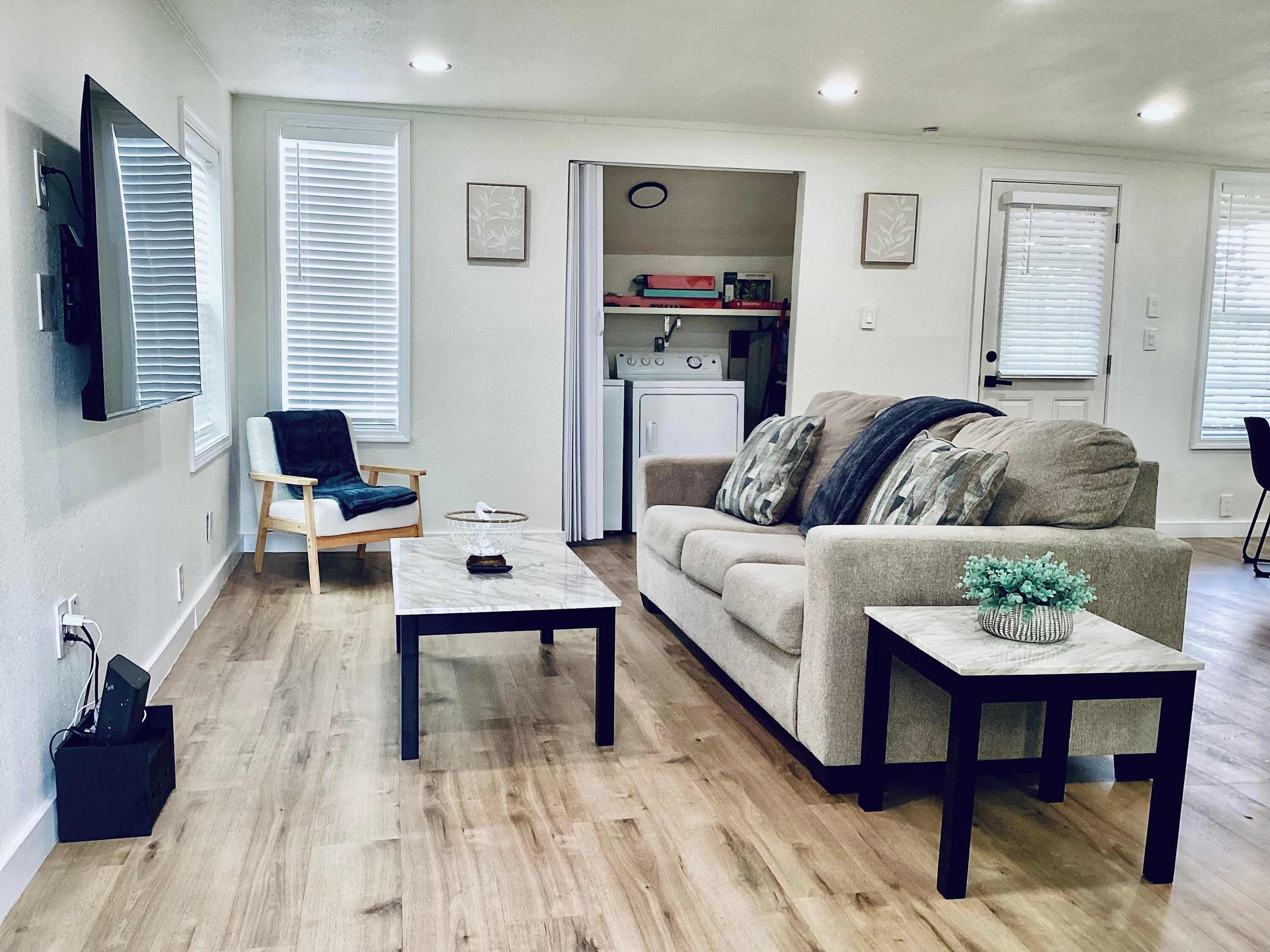 A living room with a sofa, a coffee table, a chair, and a small table with a plant. There is a kitchen area visible in the background.