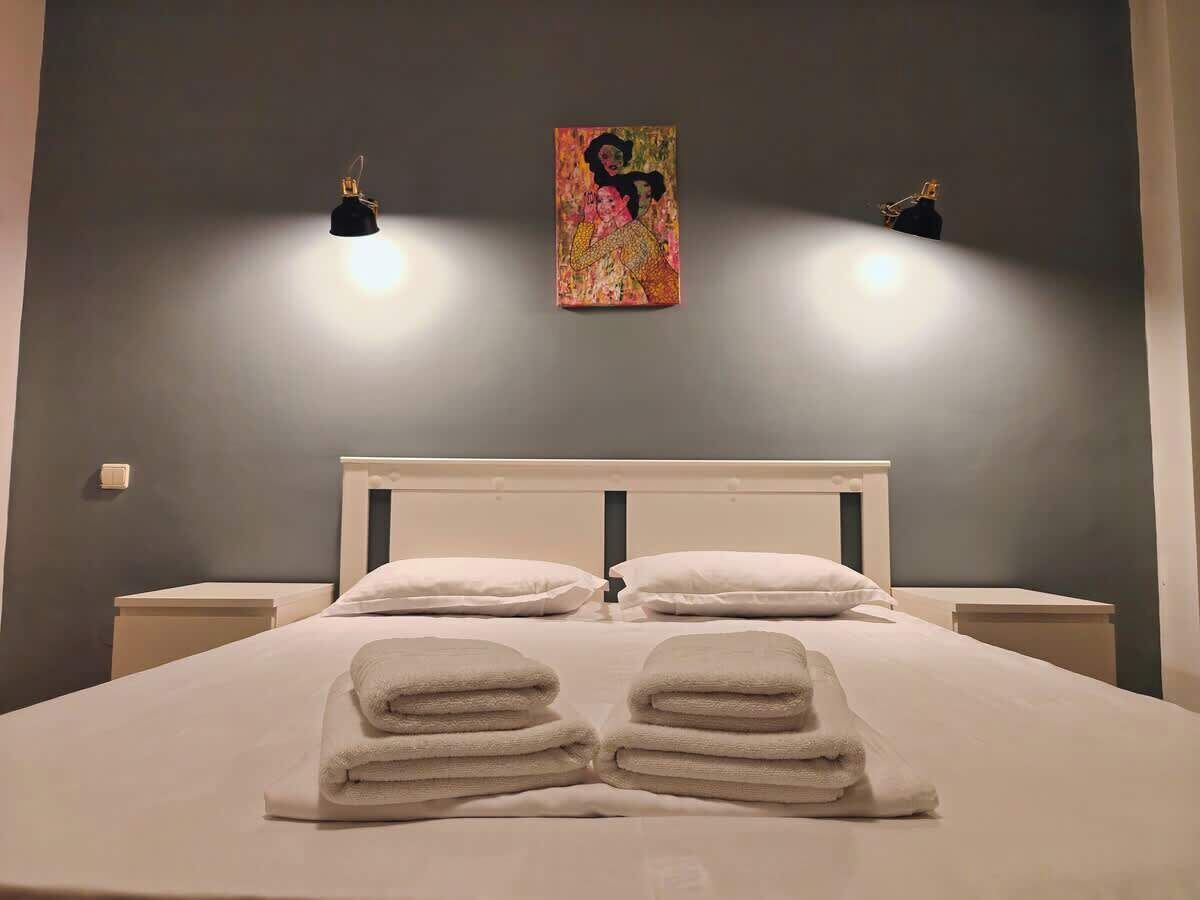 Un llit ben arropat amb tovalloles plegades al centre, dues taules de nit, un capçal i una obra d'art penjada a la paret sobre el llit.