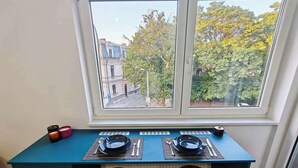 Ein Essbereich mit einem blauen Tisch, gedeckt für zwei Personen, zwei hölzernen Barhocker und einem Fenster mit Blick auf Gebäude und Bäume.