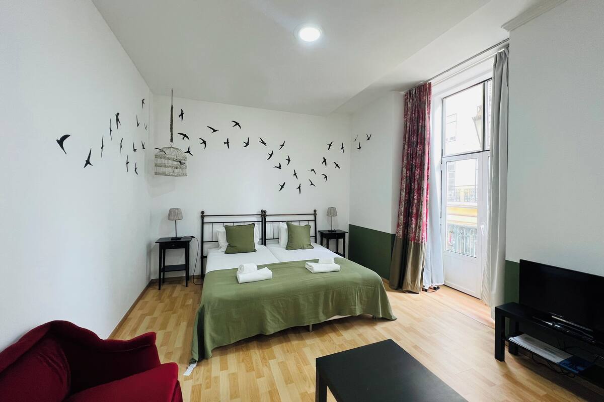 Une chambre d’hôtel avec un lit, une télévision, un canapé rouge, un plancher en bois et une fenêtre avec des rideaux.