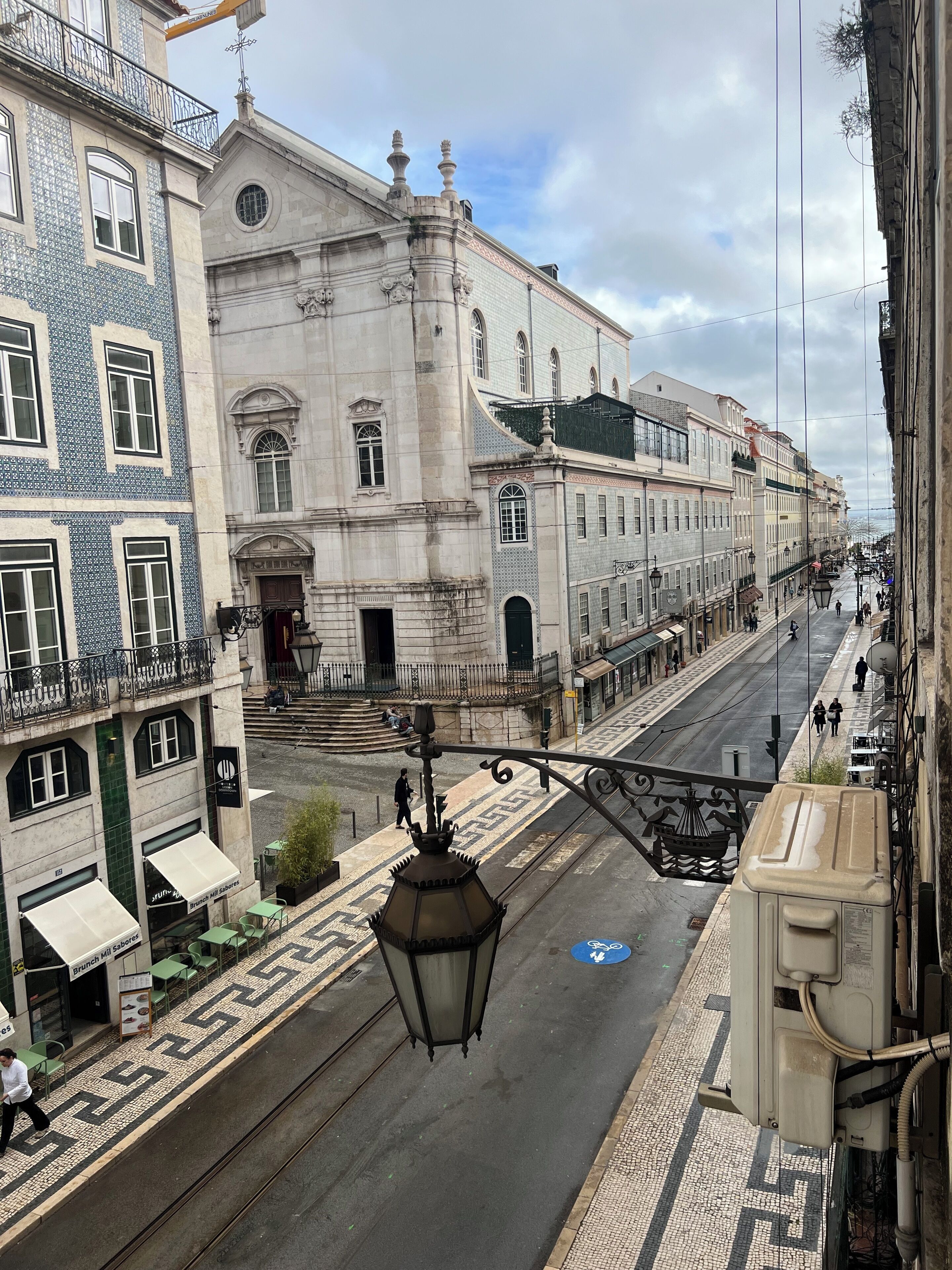 Vista da rua com edifícios com arquitetura tradicional, uma passarela para pedestres e um poste de luz.
