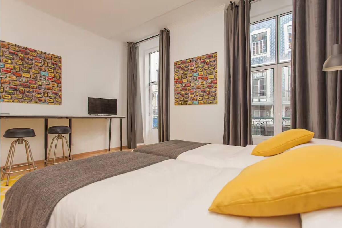Une chambre d’hôtel moderne équipée d’un lit, d’un bureau, de deux tabourets et de grandes fenêtres avec des rideaux.