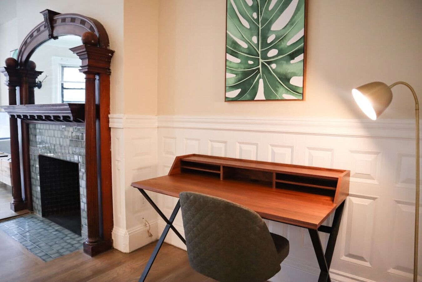 Een kamer met een houten bureau, een groene stoel, een open haard en een groot schilderij met bladmotieven aan de muur.