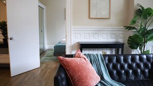 Un salon avec un canapé en cuir noir, un plaid vert et un coussin rouge. On y trouve une table basse en bois et une œuvre d’art encadrée accrochée au mur.