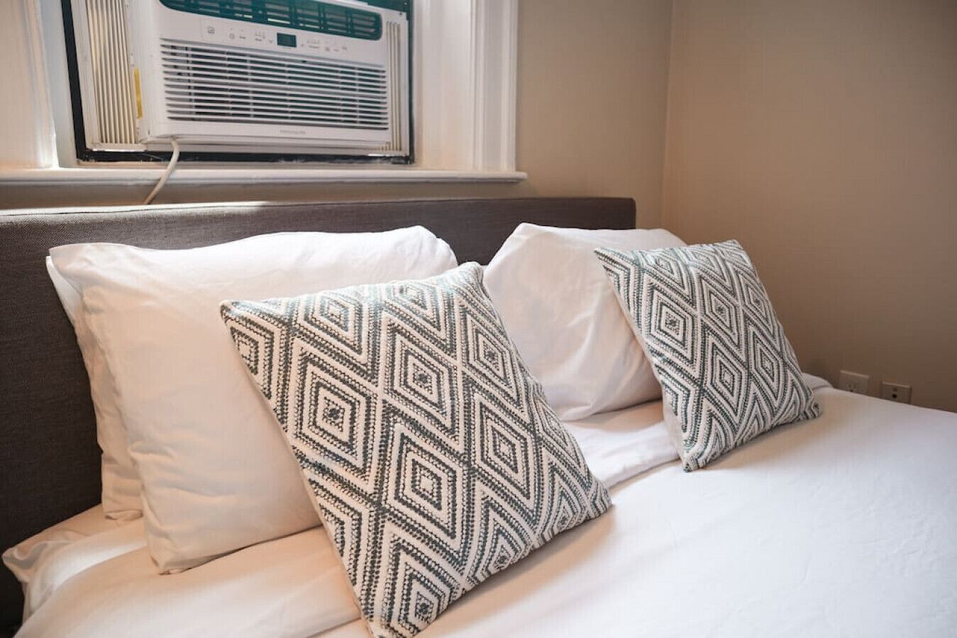 Un lit avec une literie blanche et des oreillers à motifs, un appareil de climatisation placé au-dessus de la fenêtre et un mur uni.