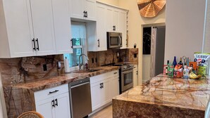 Cocina moderna con armarios blancos, encimera de granito, microondas, horno y refrigerador. Se observa una silla de mimbre y una mesa de comedor con superficie de vidrio.