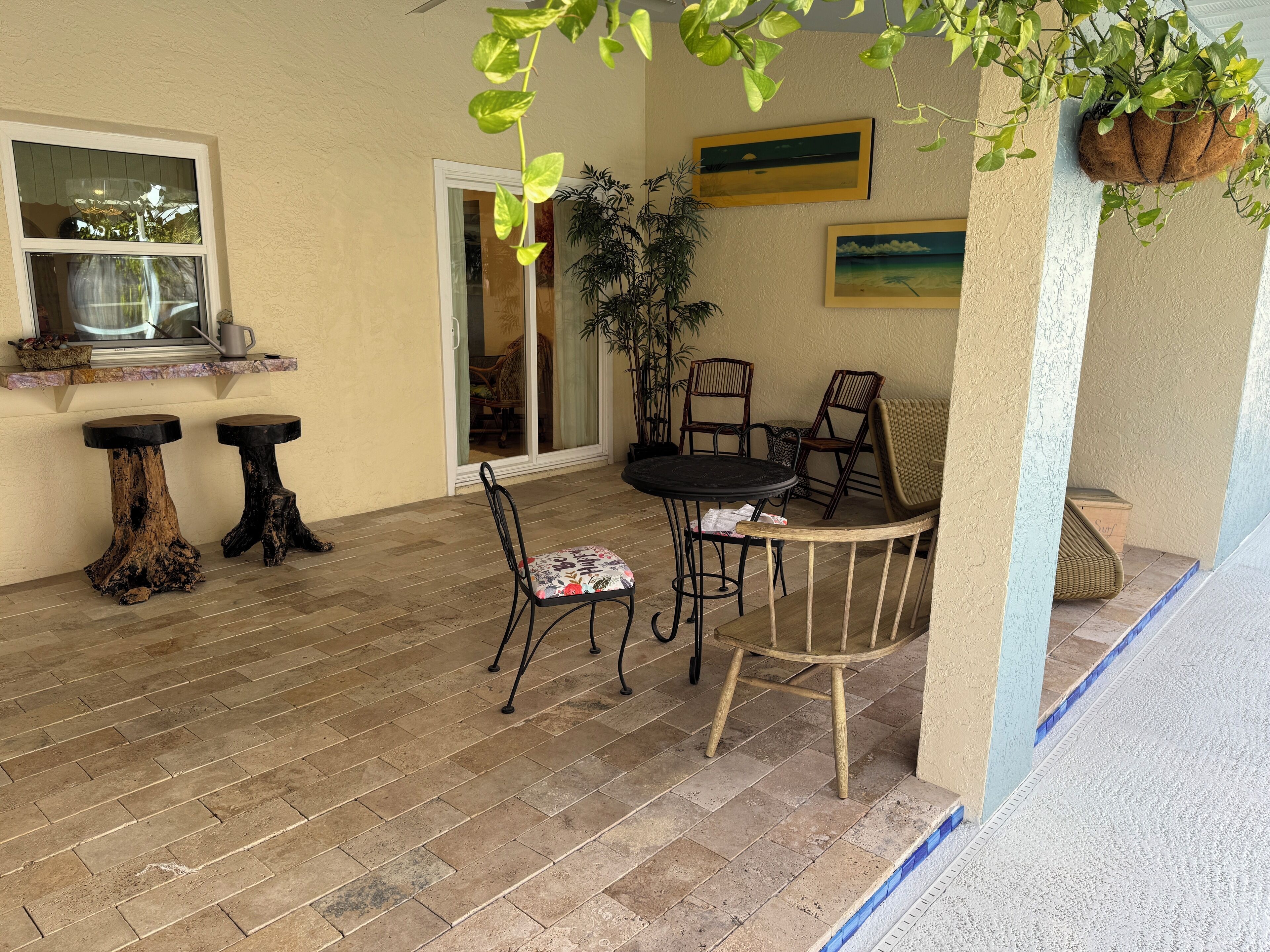 A patio with a round table and chairs, two wooden stools, a potted plant, and a window with a view.