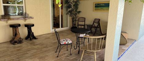 A patio with a round table and chairs, two wooden stools, a potted plant, and a window with a view.