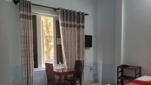Una habitación con una cama, una mesa con sillas, un ventanal con cortinas y un estante pequeño.