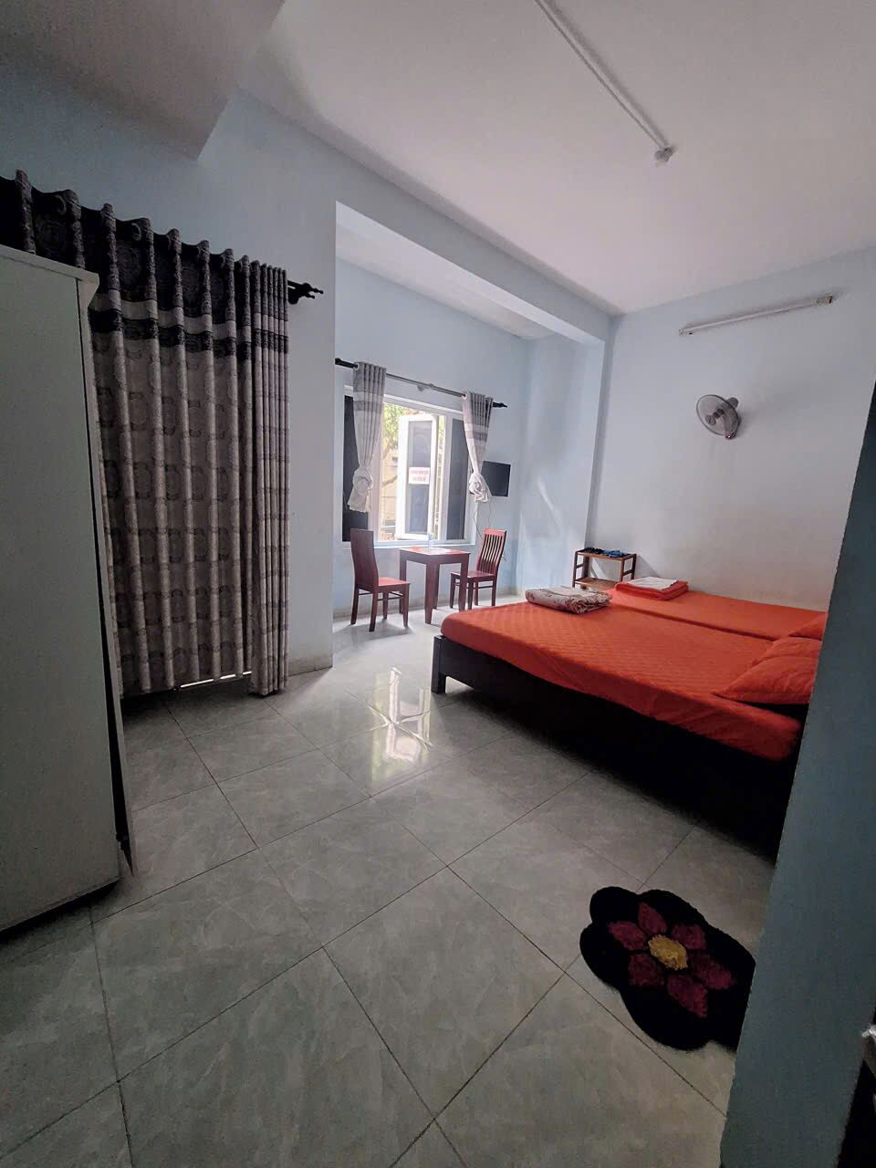 Una camera da letto con un letto, una scrivania e una sedia. Si vede una finestra con tende, un ventilatore a parete e un fiore sul pavimento.