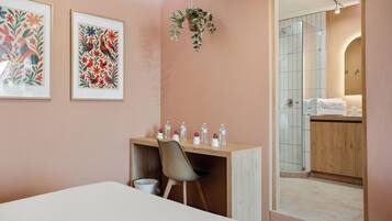 Une chambre d’hôtel avec un lit, un bureau et une chaise, une petite poubelle et une salle de bain visible à travers une porte ouverte.