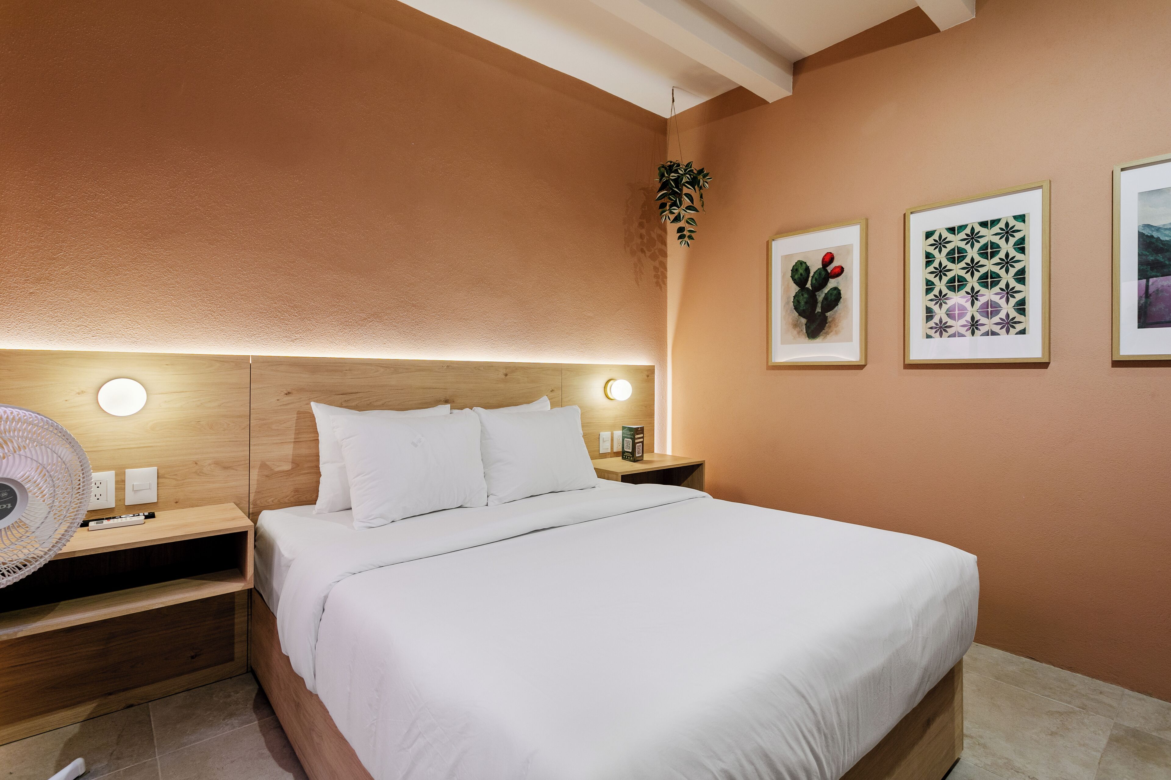 Una cama bien tendida con sábanas blancas, un cabecero de madera y una mesita de noche con una lámpara y una planta pequeña.