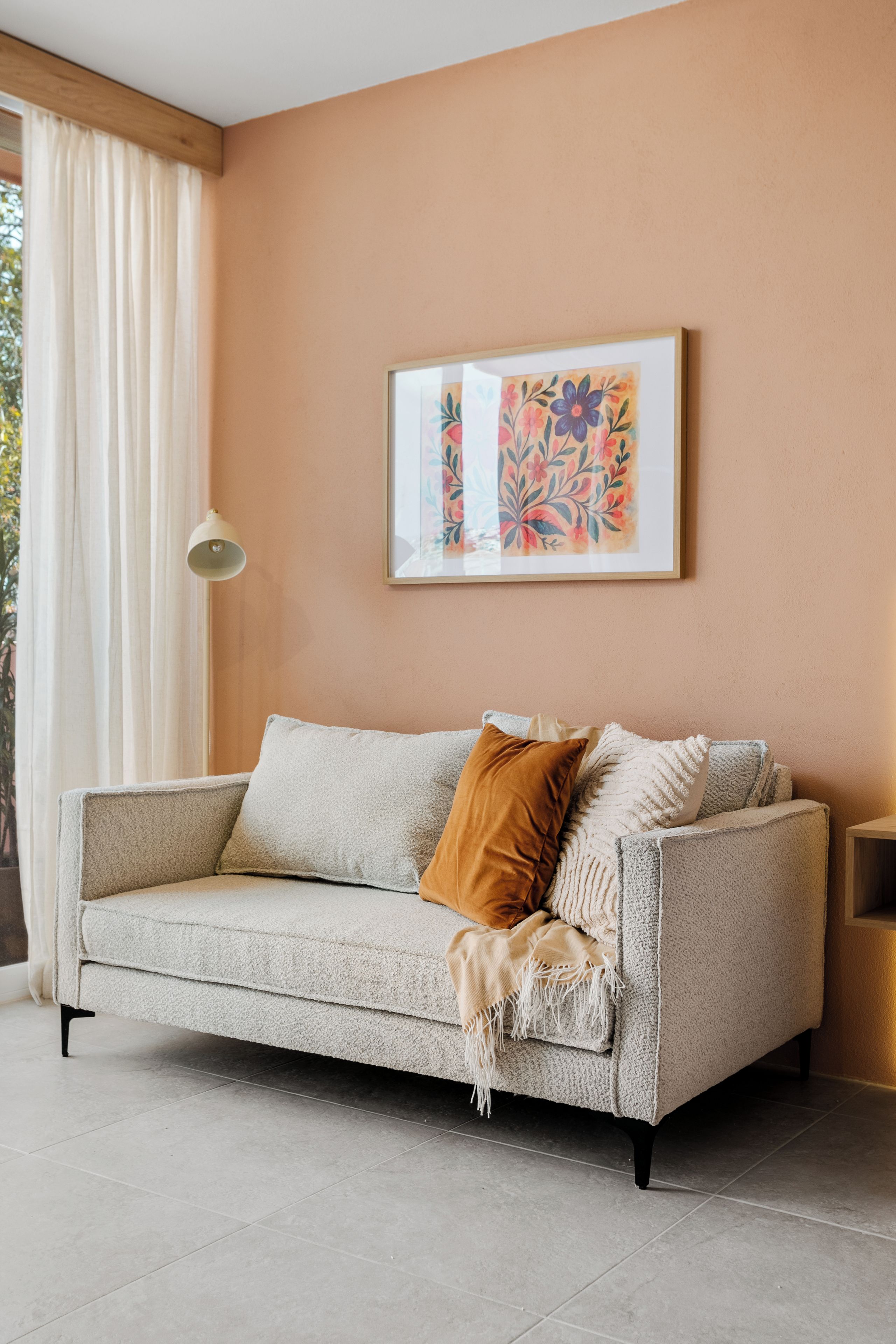 Una sala de estar con un sofá, una obra de arte enmarcada, una lámpara y una ventana con cortinas.