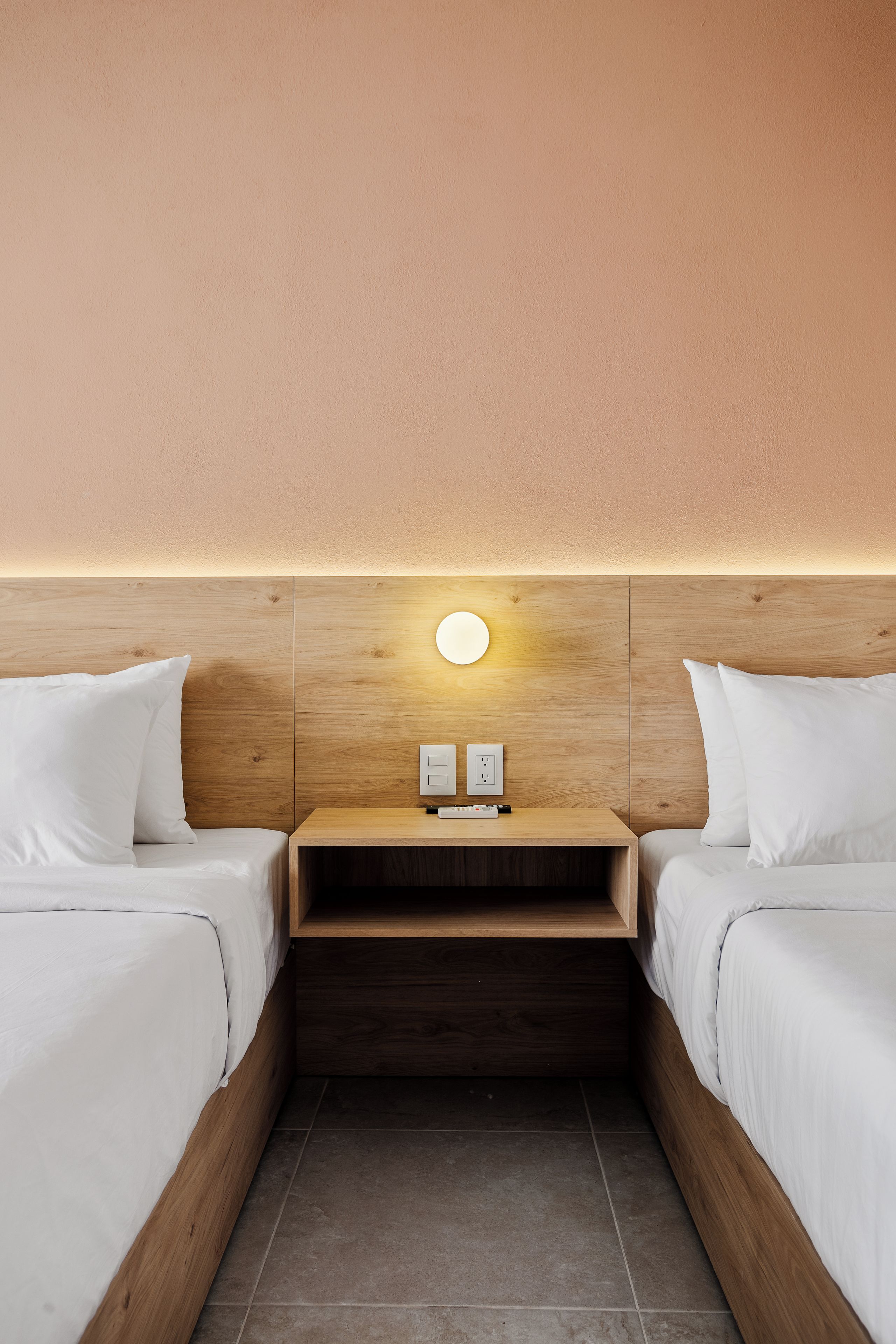 A hotel room with two beds, a wooden headboard, a nightstand with a lamp, and a wall socket.