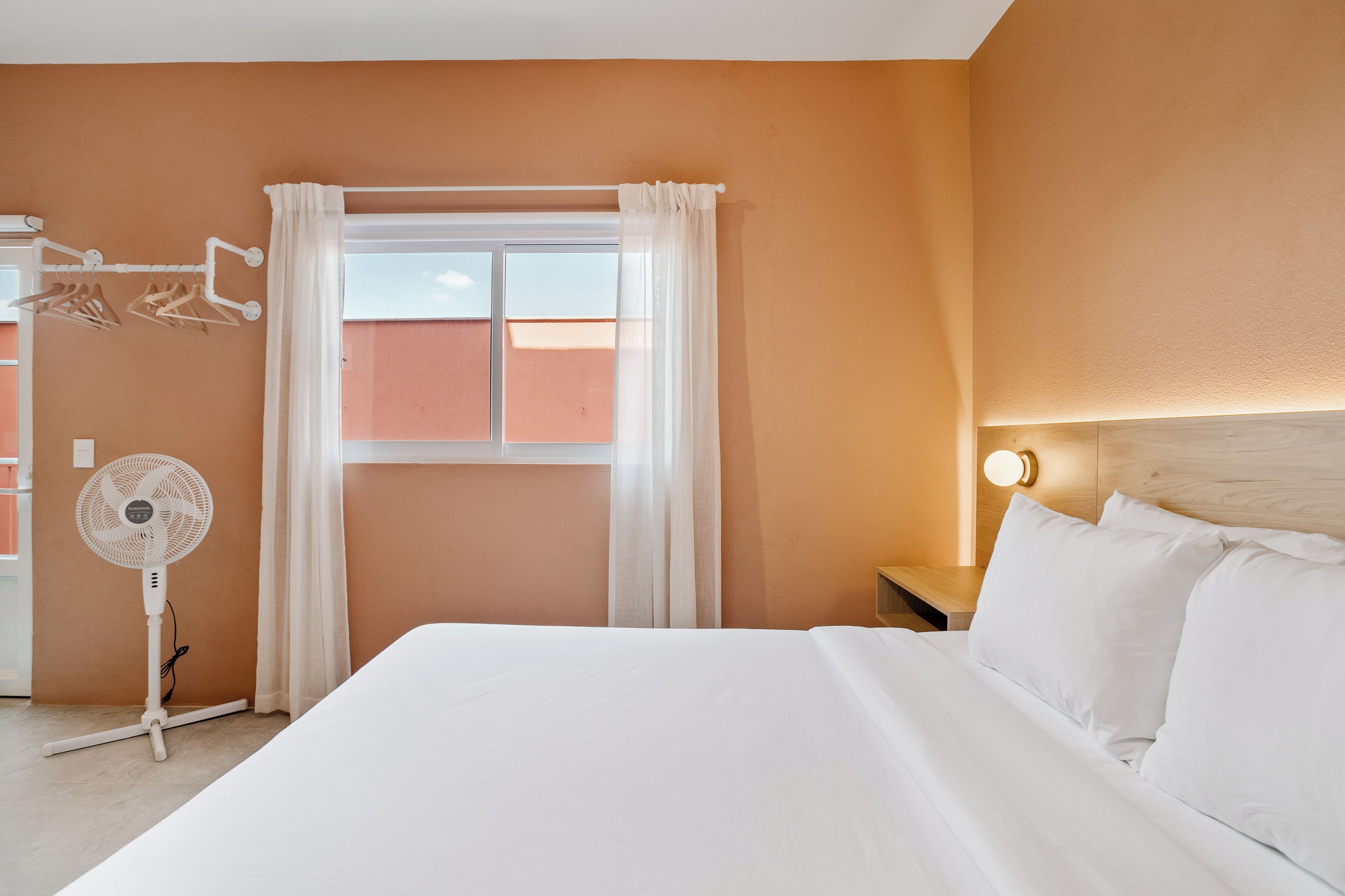 Una camera d'albergo con un letto, un comodino con lampada, una finestra con tende e un ventilatore a parete.
