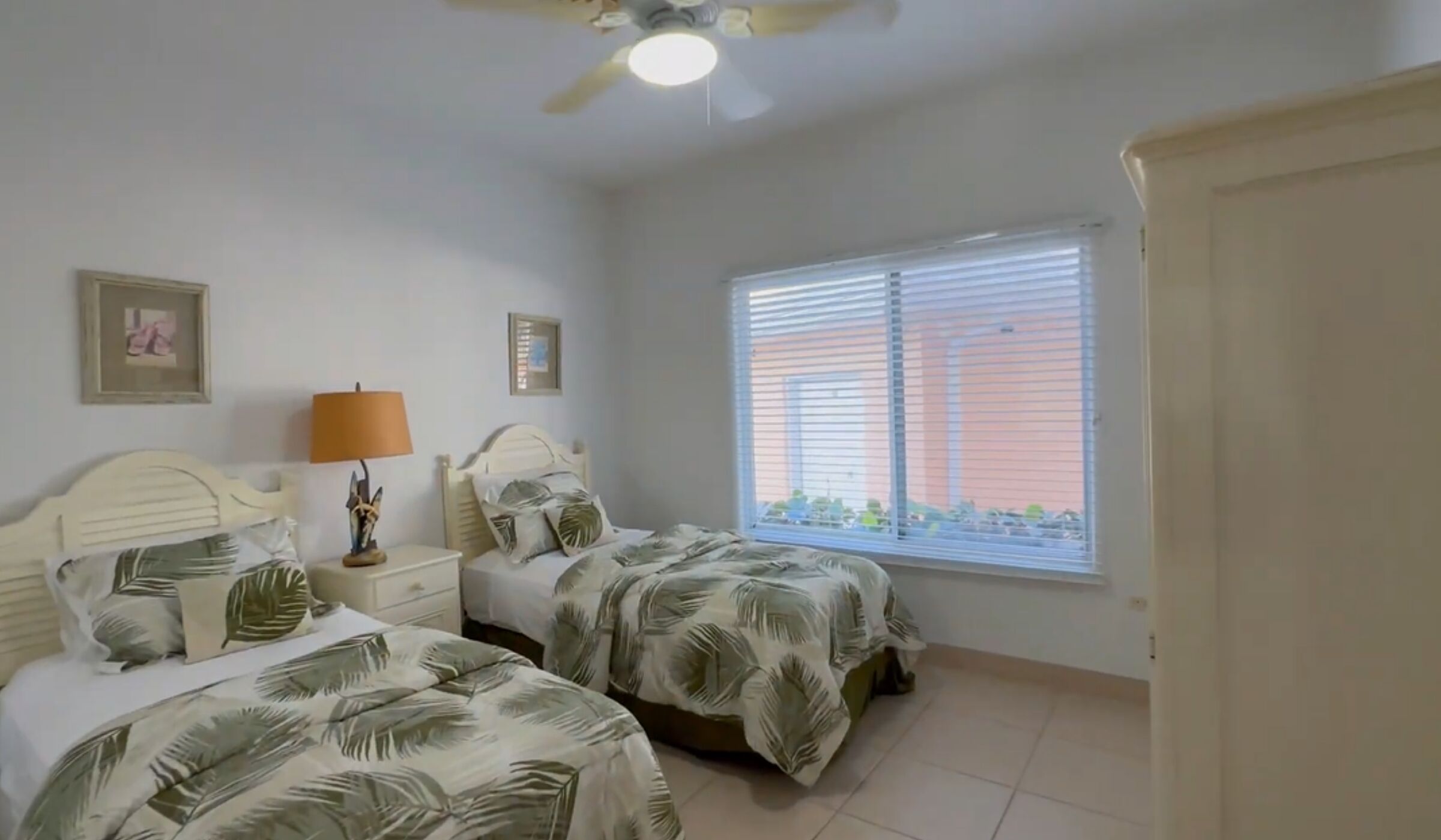 A bedroom with two beds, a ceiling fan, a window with blinds, and a framed picture on the wall.