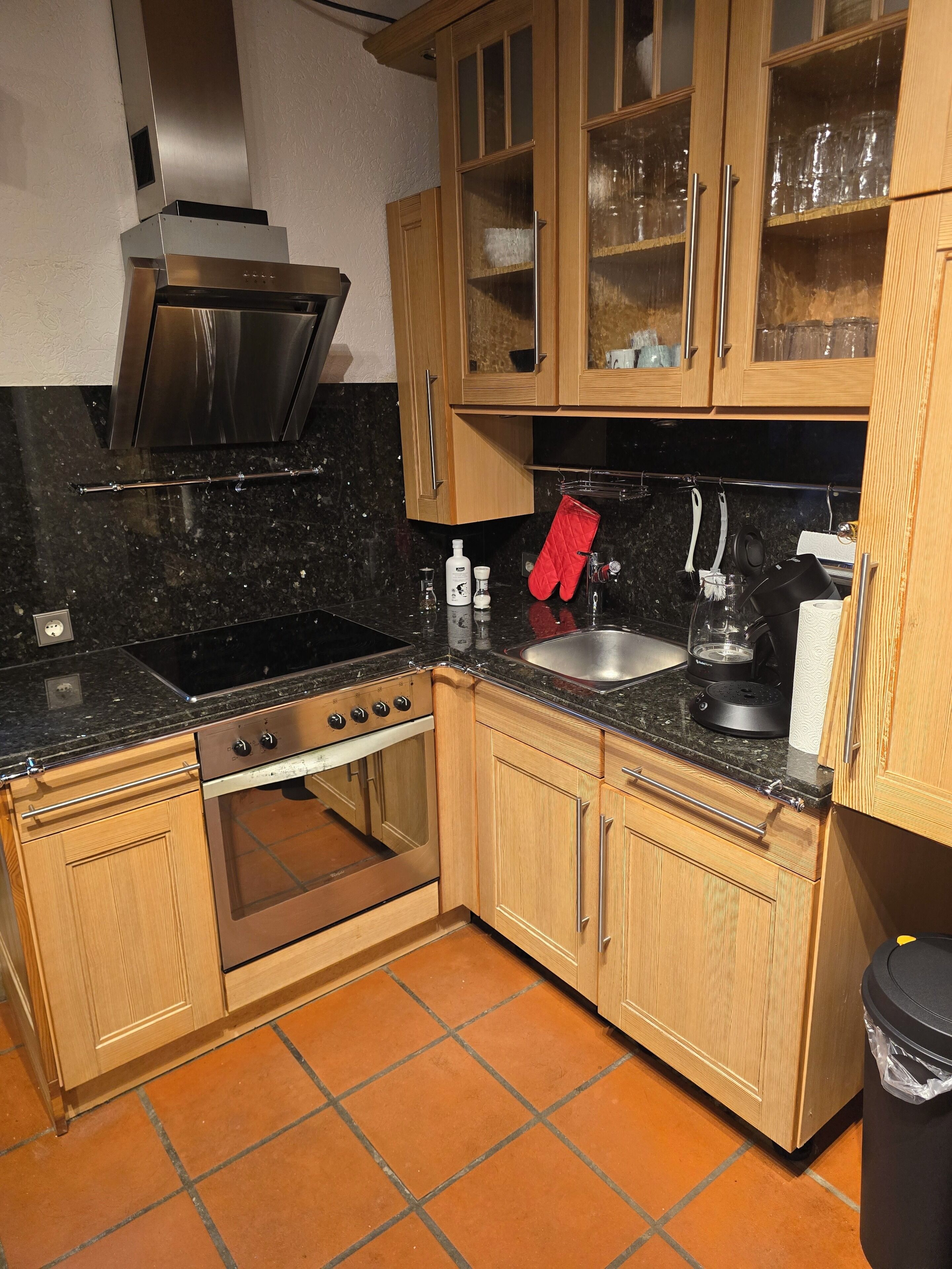 A kitchen with wooden cabinets, a stainless steel range hood, a black countertop, a sink, and a trash bin.