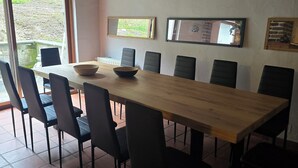 A dining area with a long wooden table, black chairs, and a bowl on the table. There are two windows with views of greenery outside.