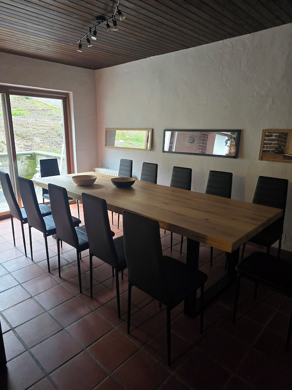 A dining area with a long wooden table, black chairs, and a bowl on the table. There are two windows with views of greenery outside.