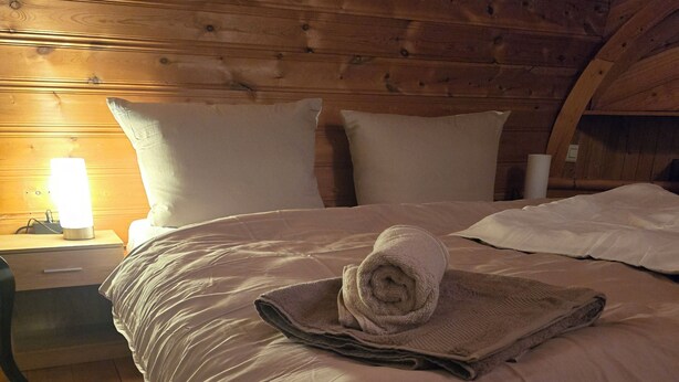 A wooden bed with a rolled towel and a blanket, a nightstand with a lamp, and a wooden floor.