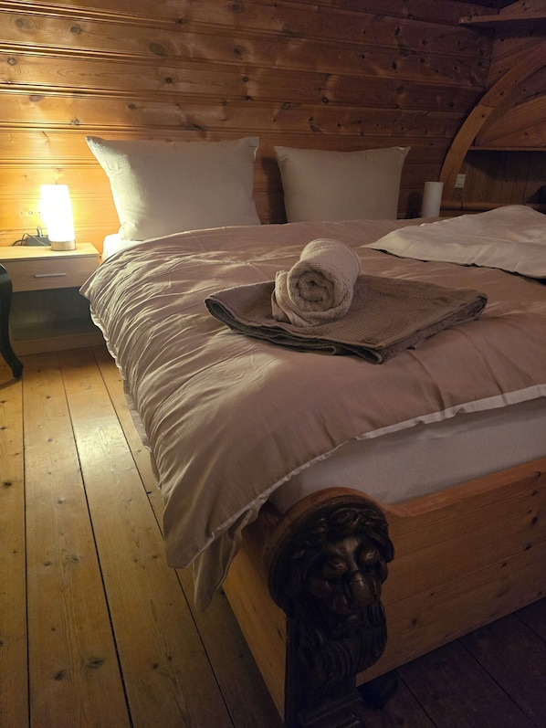 Una cama de madera con una toalla enrollada y una manta, una mesita de noche con una lámpara y un suelo de madera.