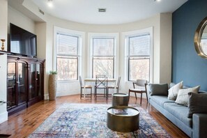 A living room with a wooden floor, a blue sofa, a dining table with chairs, and three windows with blinds. - Newly renovated in a prime location in Kenmore Square.  Walking distance to Downtown + Universities (Boston)