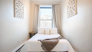 Ein schmales Hotelzimmer mit Bett, einem kleinen Tisch und einem Fenster mit Vorhängen.