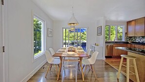 Een moderne keuken met houten kastjes, een eethoek met een lange tafel en stoelen, en grote ramen met uitzicht op groen.