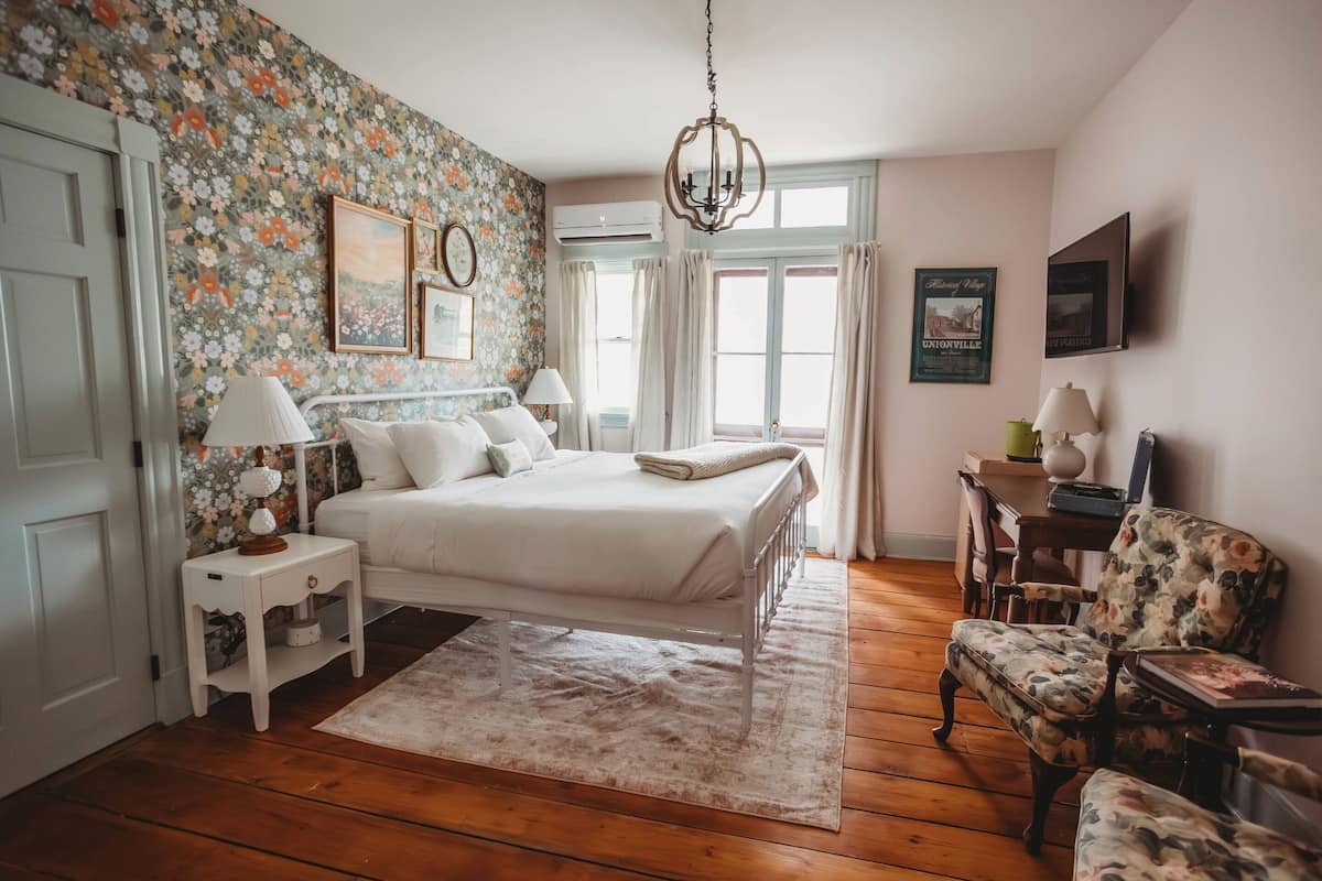 A bedroom with a bed, bedside tables, a chair, a chandelier, and floral wallpaper.