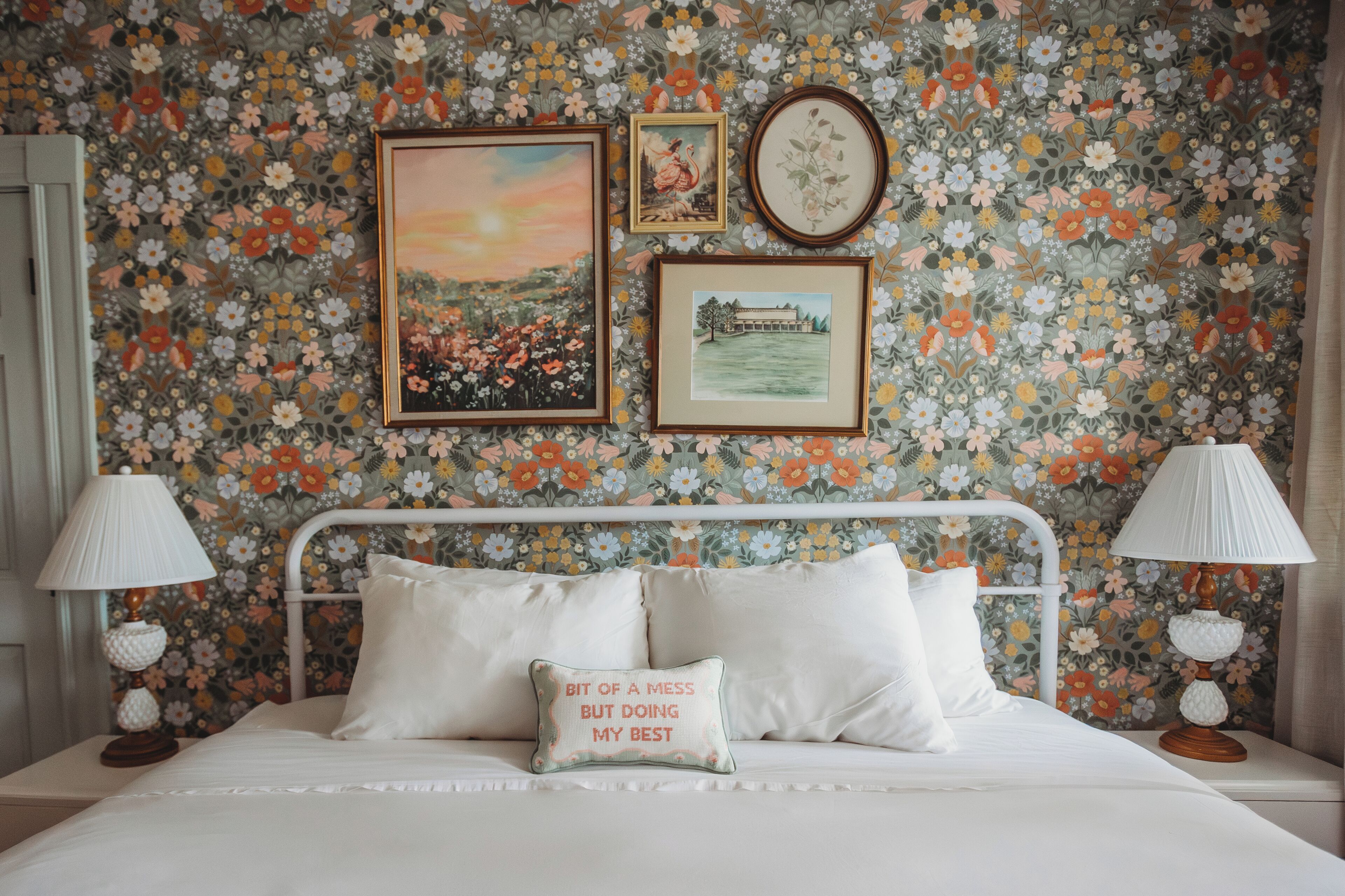 A bed with white bedding, a pillow with text, and a floral wallpaper.