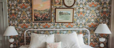 A bed with white bedding, a pillow with text, and a floral wallpaper.