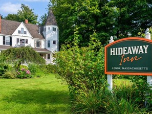 A sign for Hideaway Inn in Lenox, Massachusetts, with a white building and a tower in the background. - Hideaway Inn Lenox (Lenox)