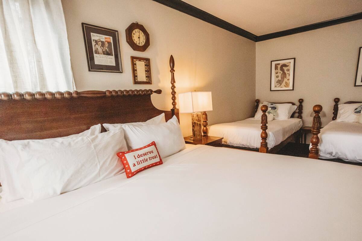 A hotel room with a large bed, two smaller beds, a wooden headboard, a bedside table with a lamp, and wall-mounted clocks and pictures.