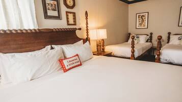 A hotel room with a large bed, two smaller beds, a wooden headboard, a bedside table with a lamp, and wall-mounted clocks and pictures.