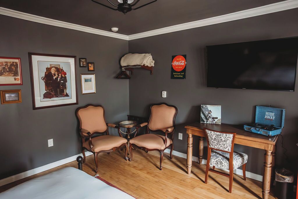 A room with a bed, two chairs, a small table, a television, and framed pictures on the wall.