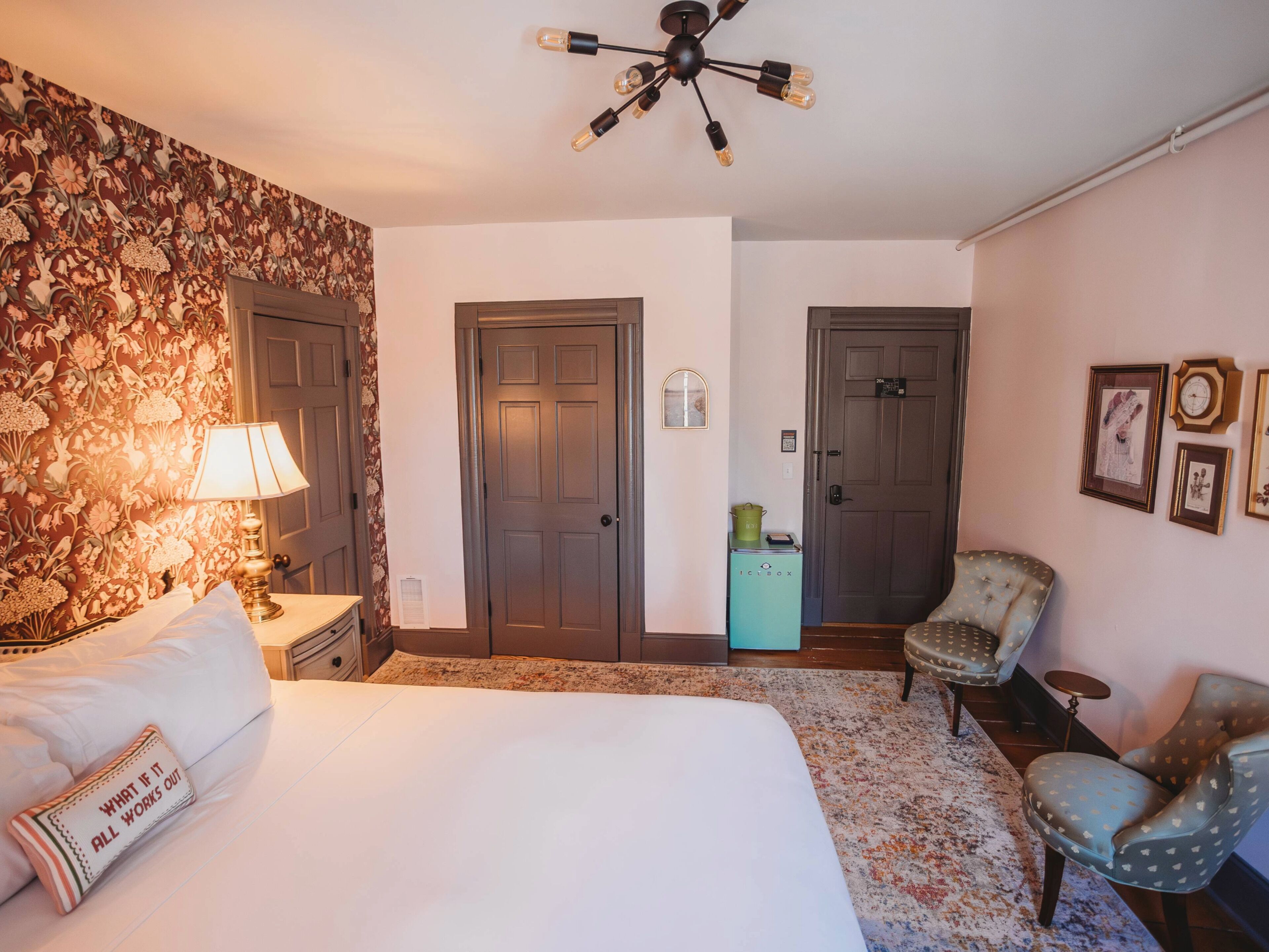 Une chambre d’hôtel avec un lit, deux portes, une lampe, une chaise, une petite table et un ventilateur de plafond.