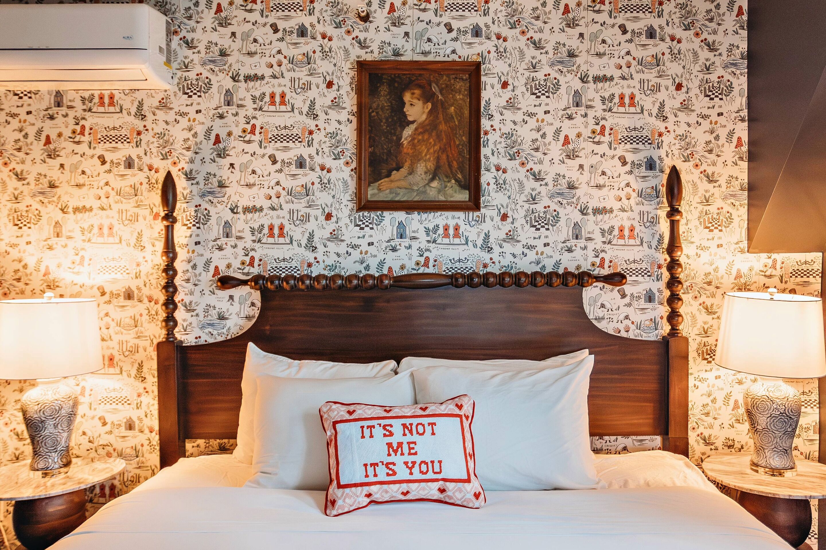 A hotel room with a patterned wallpaper, a wooden headboard, a bed with white linens, and a decorative pillow with text.