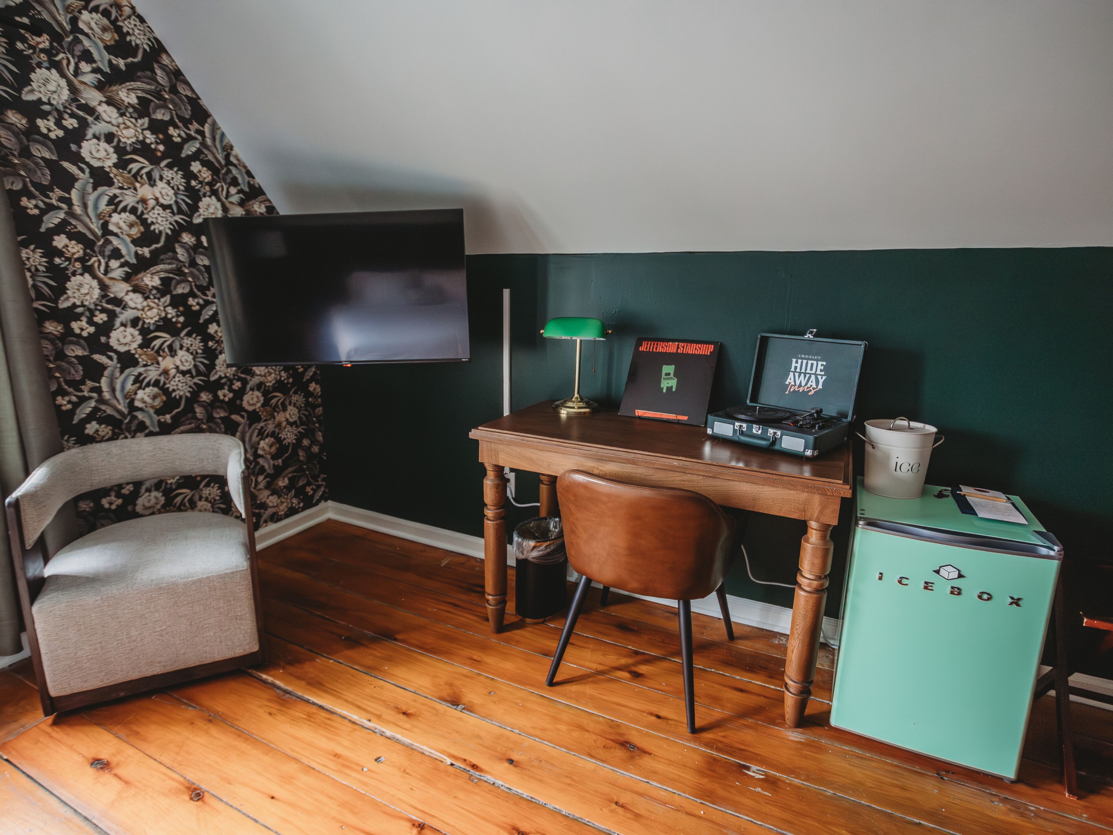 A room with a wooden desk, a chair, a television mounted on the wall, and a vintage icebox.