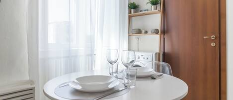 Una mesa redonda para comer con platos blancos, cubiertos y vasos, frente a una pared blanca con una puerta de madera y una repisa con objetos decorativos.