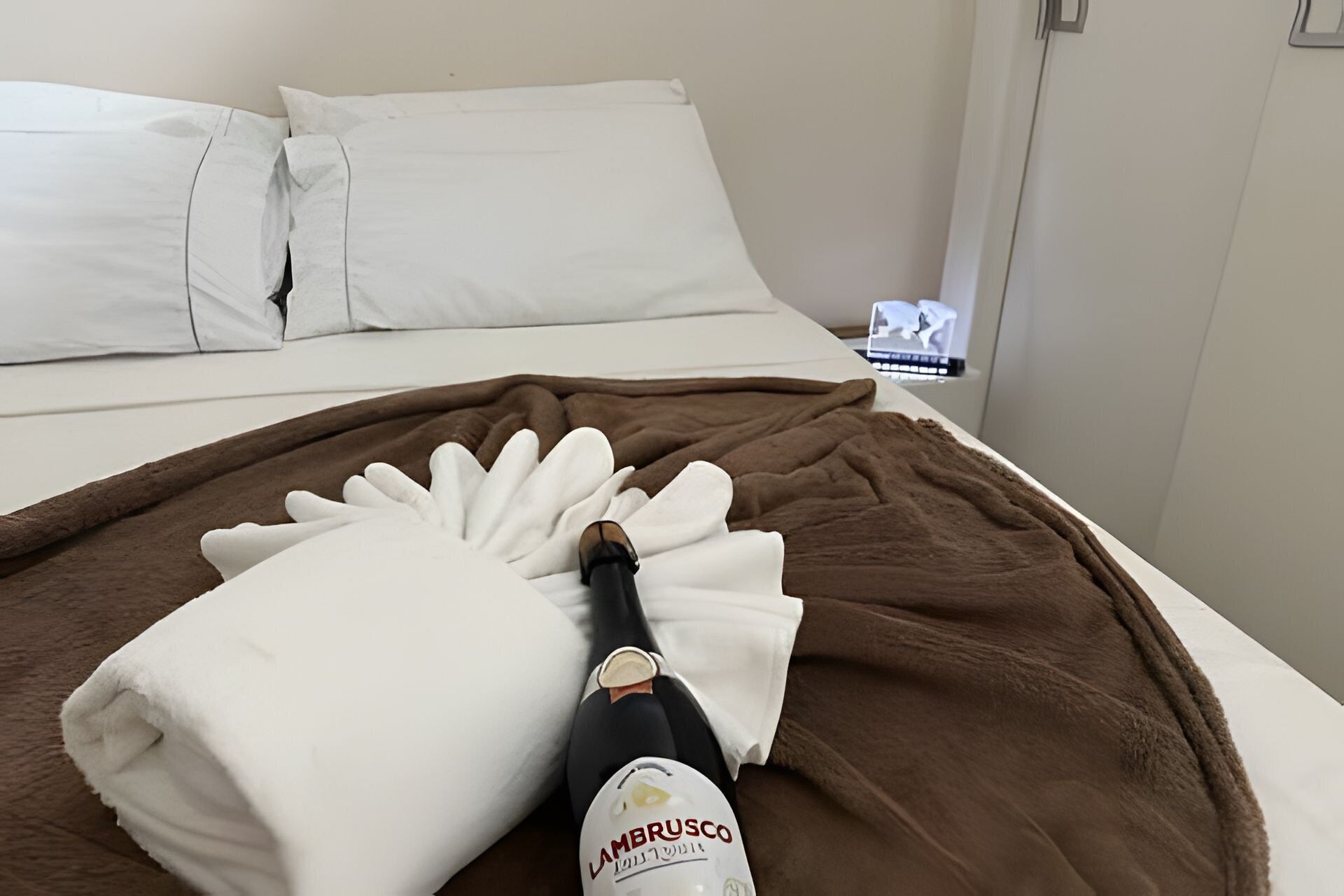 Una cama con una manta marrón, una almohada blanca y una botella de vino Amarusco.