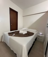 Una cama bien hecha con una colcha blanca, un edredón marrón y una almohada blanca. Se ve una puerta de madera y una mesita de noche con una lámpara y una caja de pañuelos.