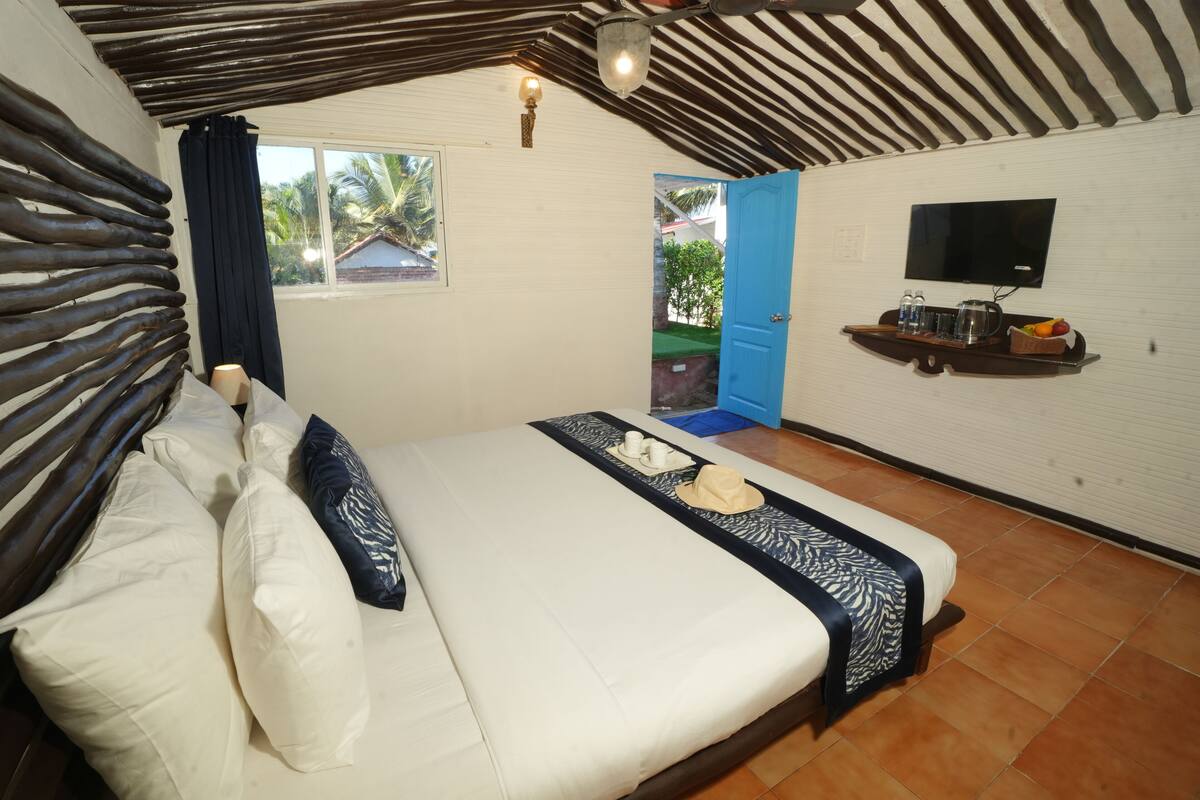 A bedroom with a bed, pillows, a bedside table with a TV, and a window with a view of palm trees.