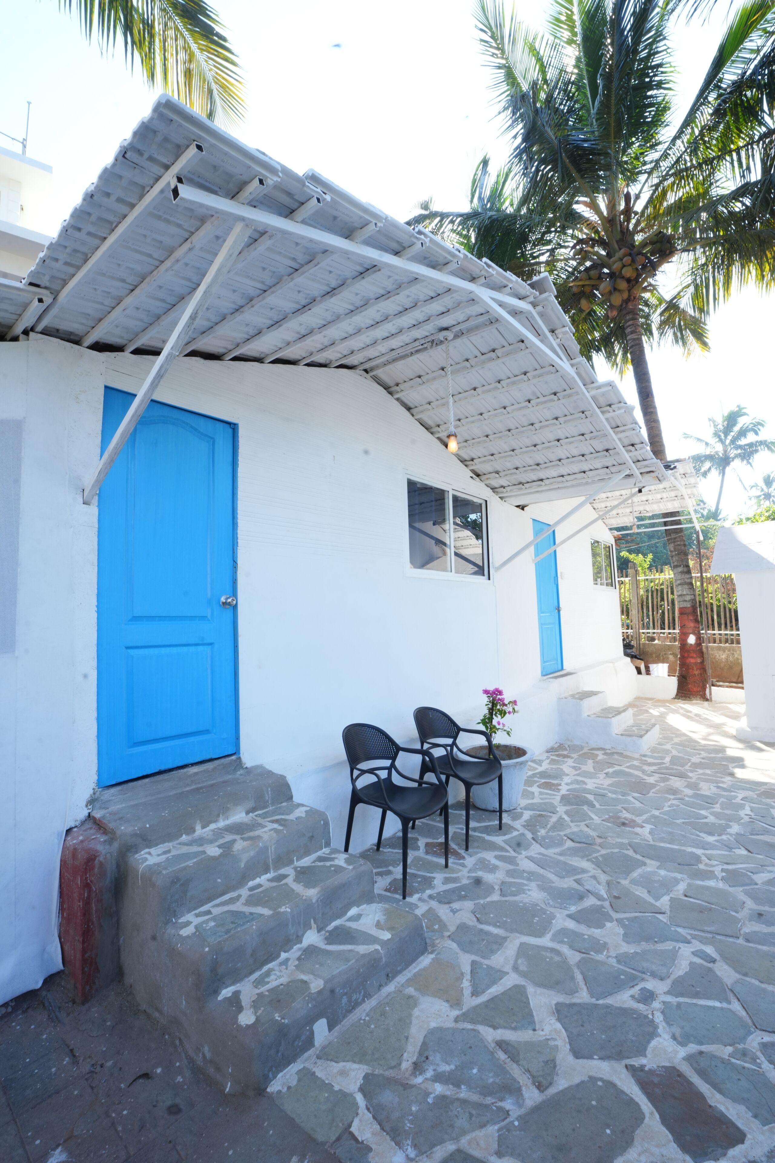 Un edificio blanco con puerta azul y un porche cubierto con suelo de piedra y dos sillas.