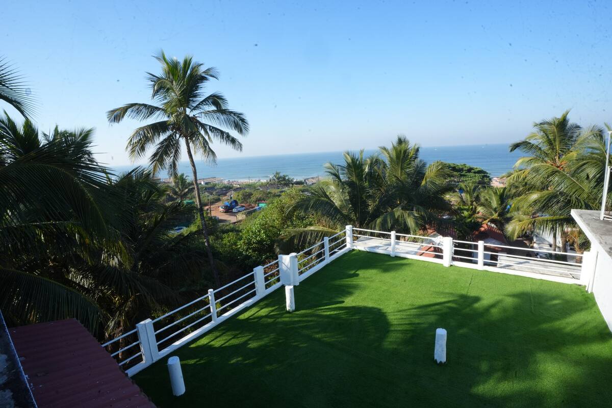 A view from a balcony with a white railing, overlooking a green lawn and a beach with palm trees.