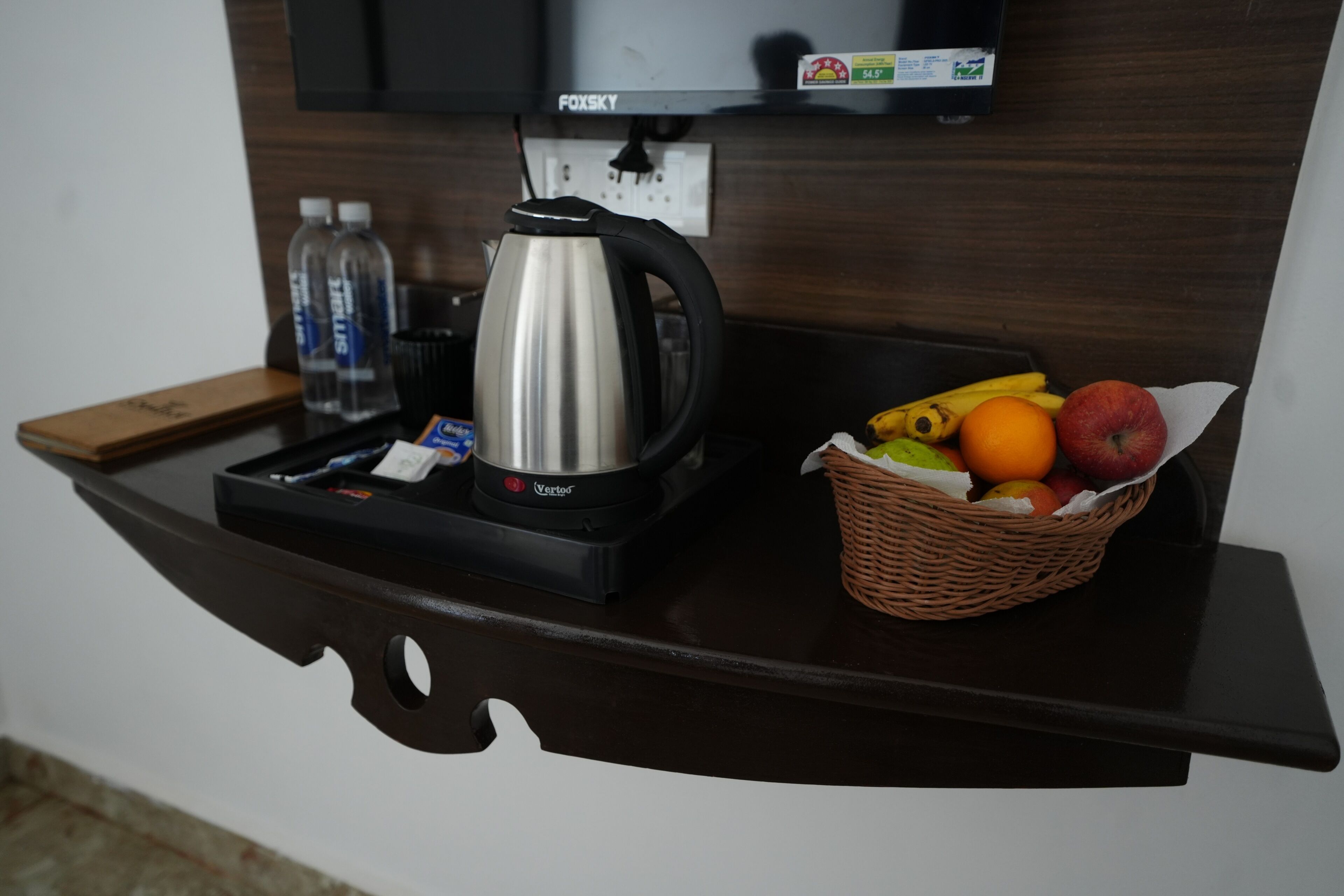 A kitchen counter with a coffee maker, water bottles, a basket of fruit, and a television mounted on the wall.