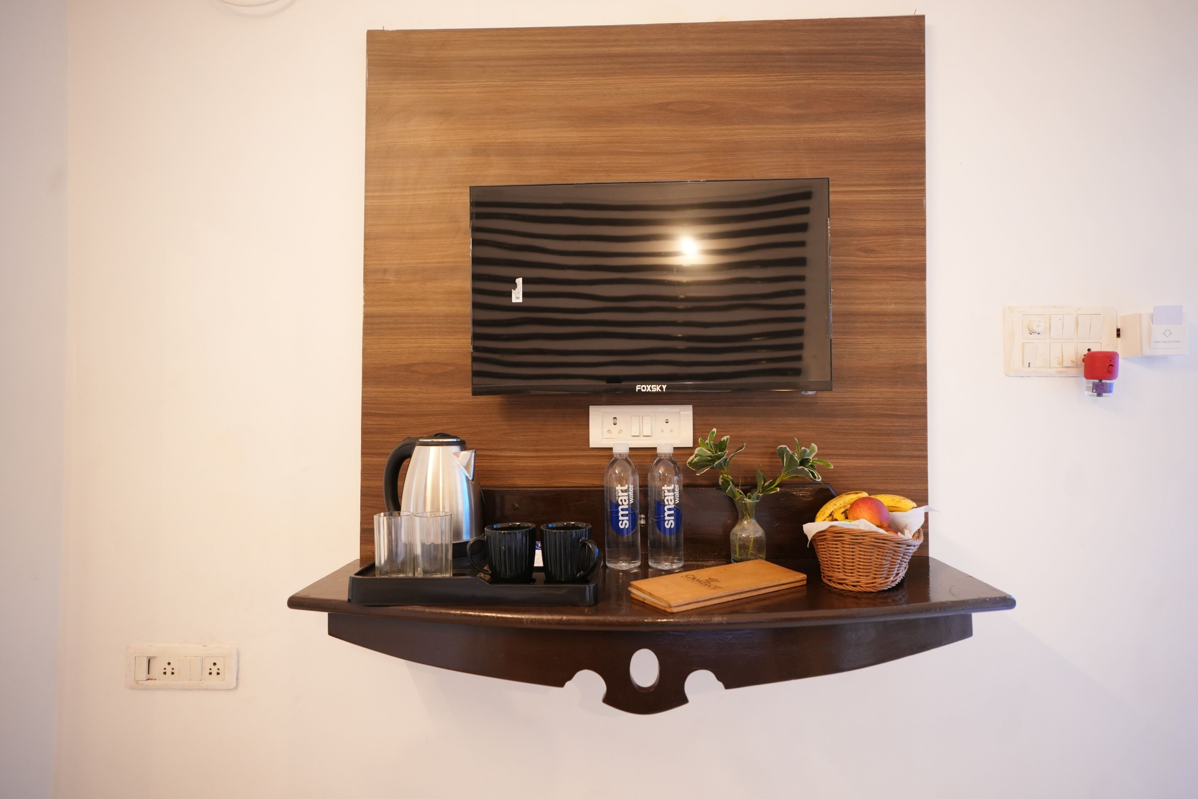 A modern living area with a flat-screen TV mounted on a wooden panel, a coffee station with a kettle, mugs, and a basket of fruit.