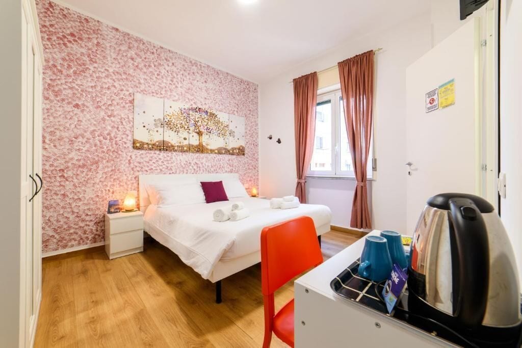 A bedroom with a bed, a nightstand, a desk with a kettle, and a window with curtains.