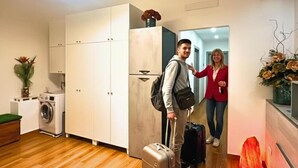Une chambre d’hôtel moderne avec un plancher en bois, un lave-linge, une grande armoire et un petit coin salon agrémenté d’un coussin vert.