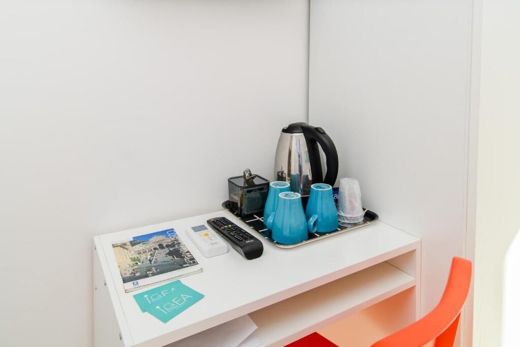 A hotel room with a white desk, a kettle, a tray with blue cups, a remote control, and a postcard.